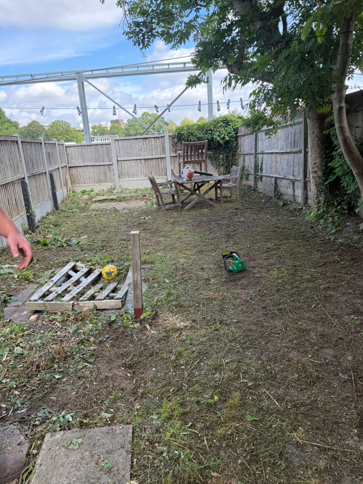 Back garden mid-clearance with cut grass on the lawn, a green chainsaw on the ground, and a leaf blower on a wooden table beside timber fencing