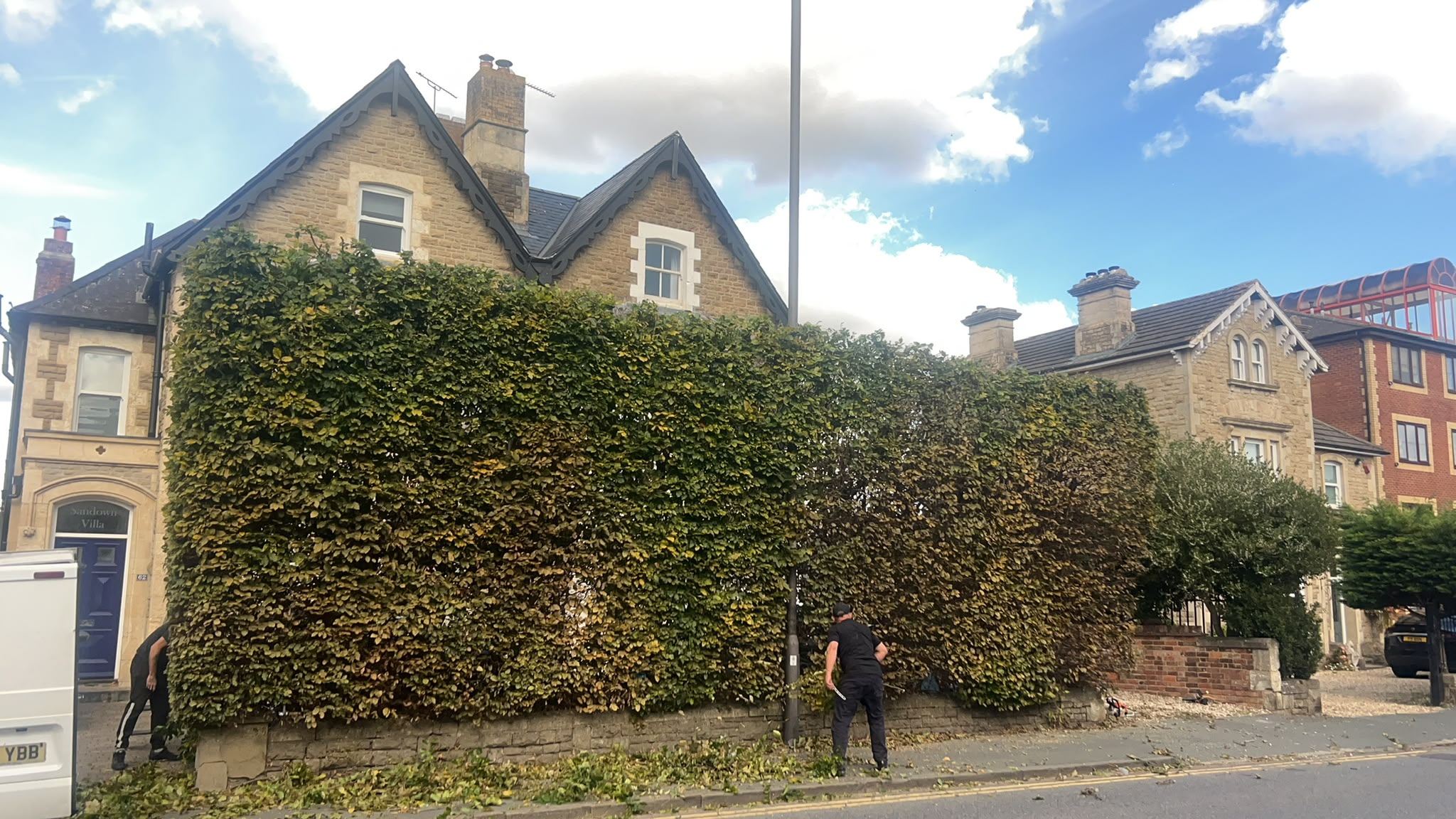 Workers using long-reach trimmers on a tall hedge in front of a stone house with clippings on the pavement