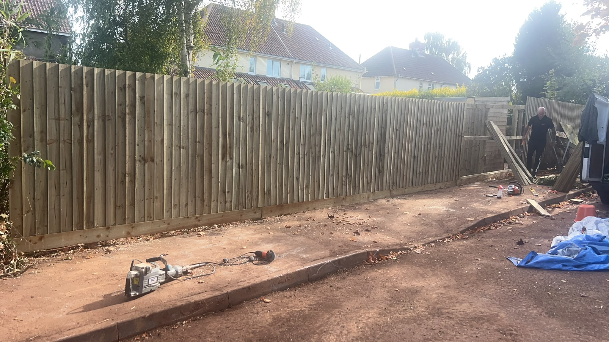 Long newly built wooden closeboard fence with a jackhammer in the foreground and a worker near a white van