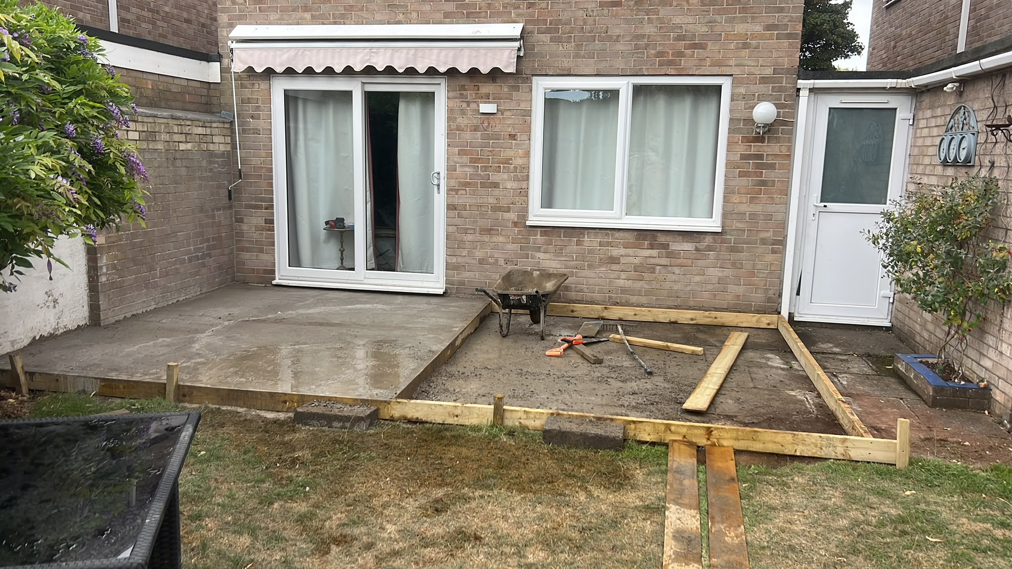 Timber formwork for a new patio half-filled with wet concrete beside a brick house, with a wheelbarrow and shovel
