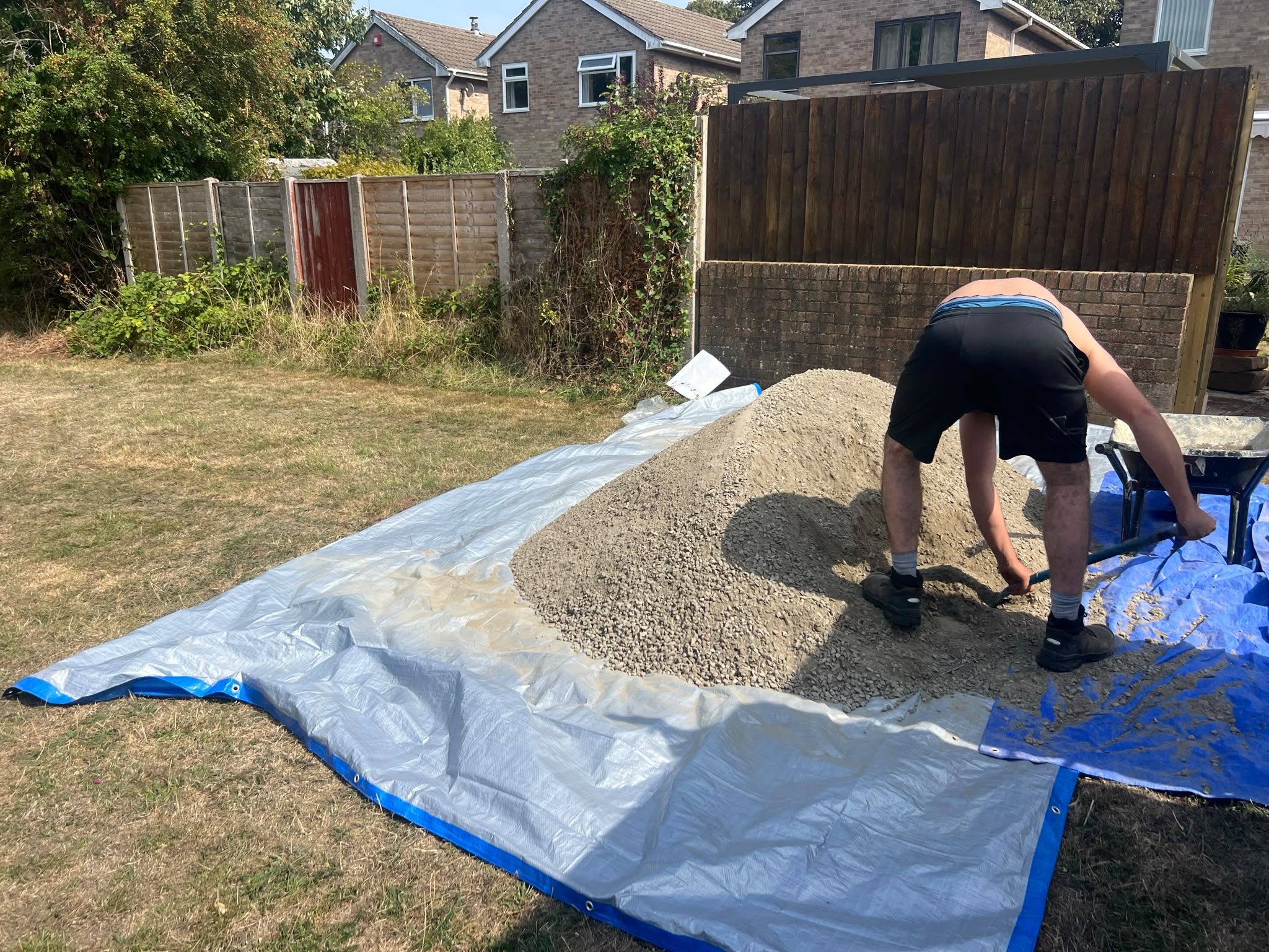 Worker shovelling grey aggregate from a tarp into a blue wheelbarrow in a sunny residential garden