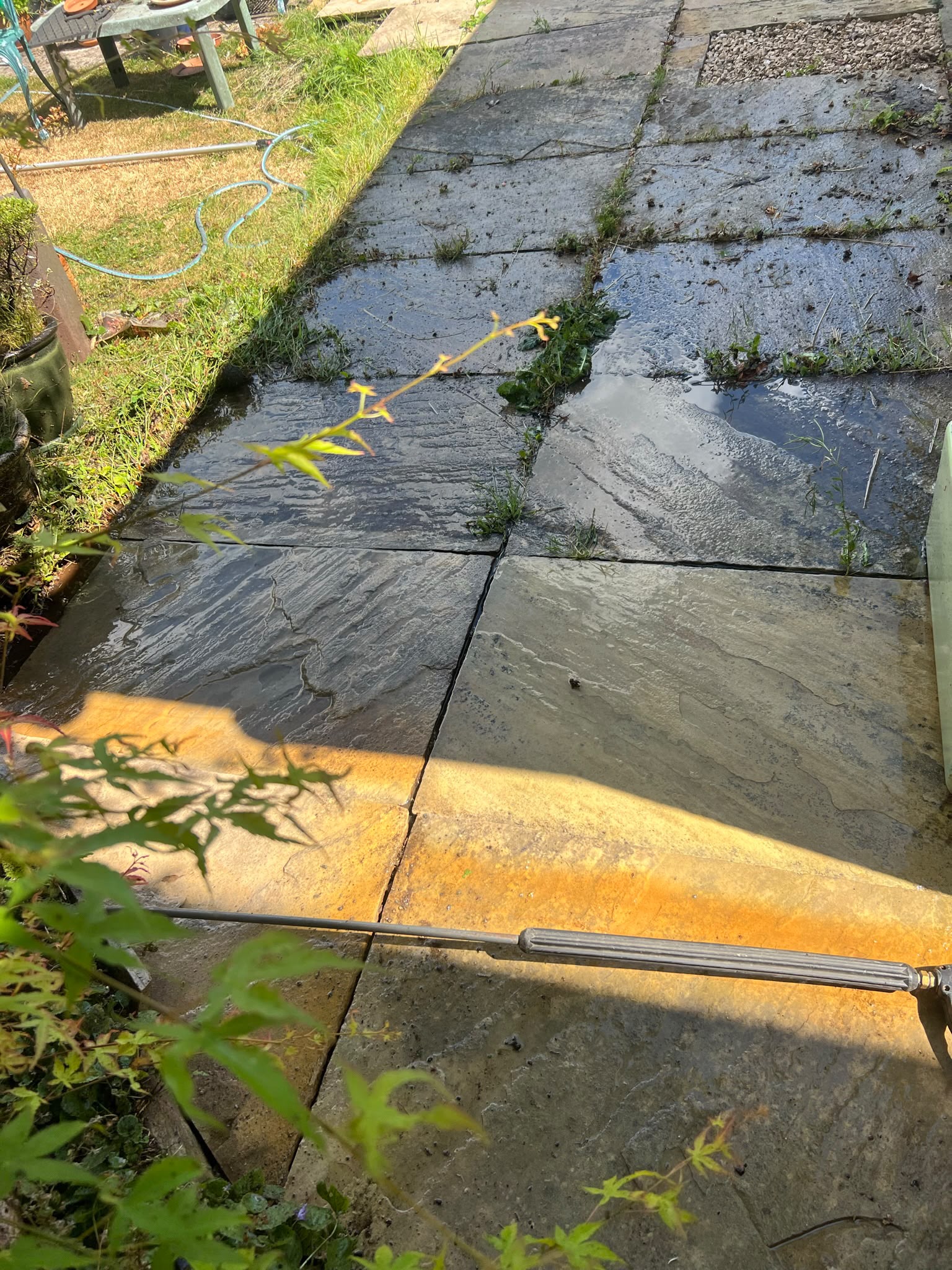 Large stone patio slabs showing dirty mossy areas beside freshly cleaned sandy-yellow stone with a pressure washer wand