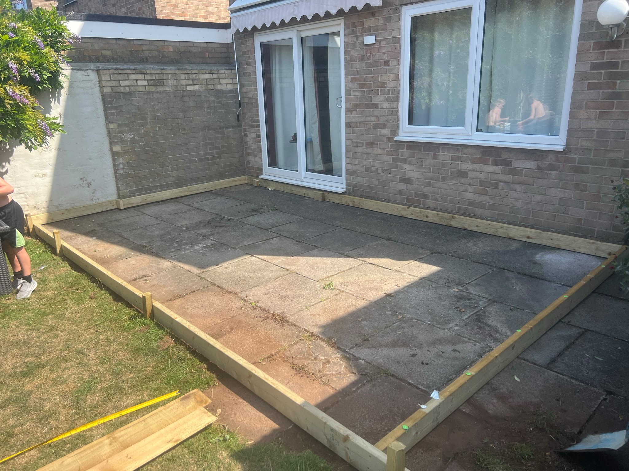 Rectangular timber frame being built over grey paving slabs next to a brick house with sliding doors