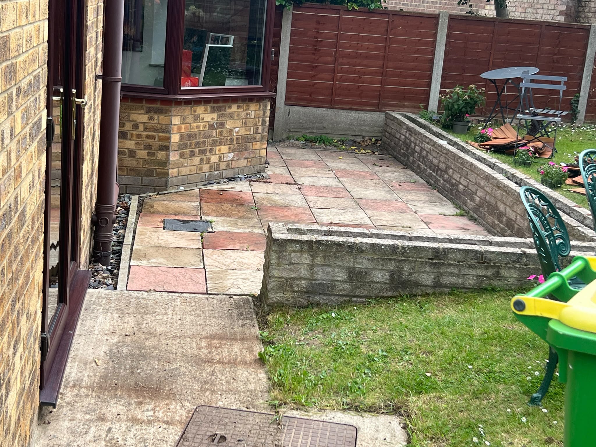 Multi-coloured stone patio slabs with low brick retaining borders, lawn, and wooden fence beside a yellow-brick house