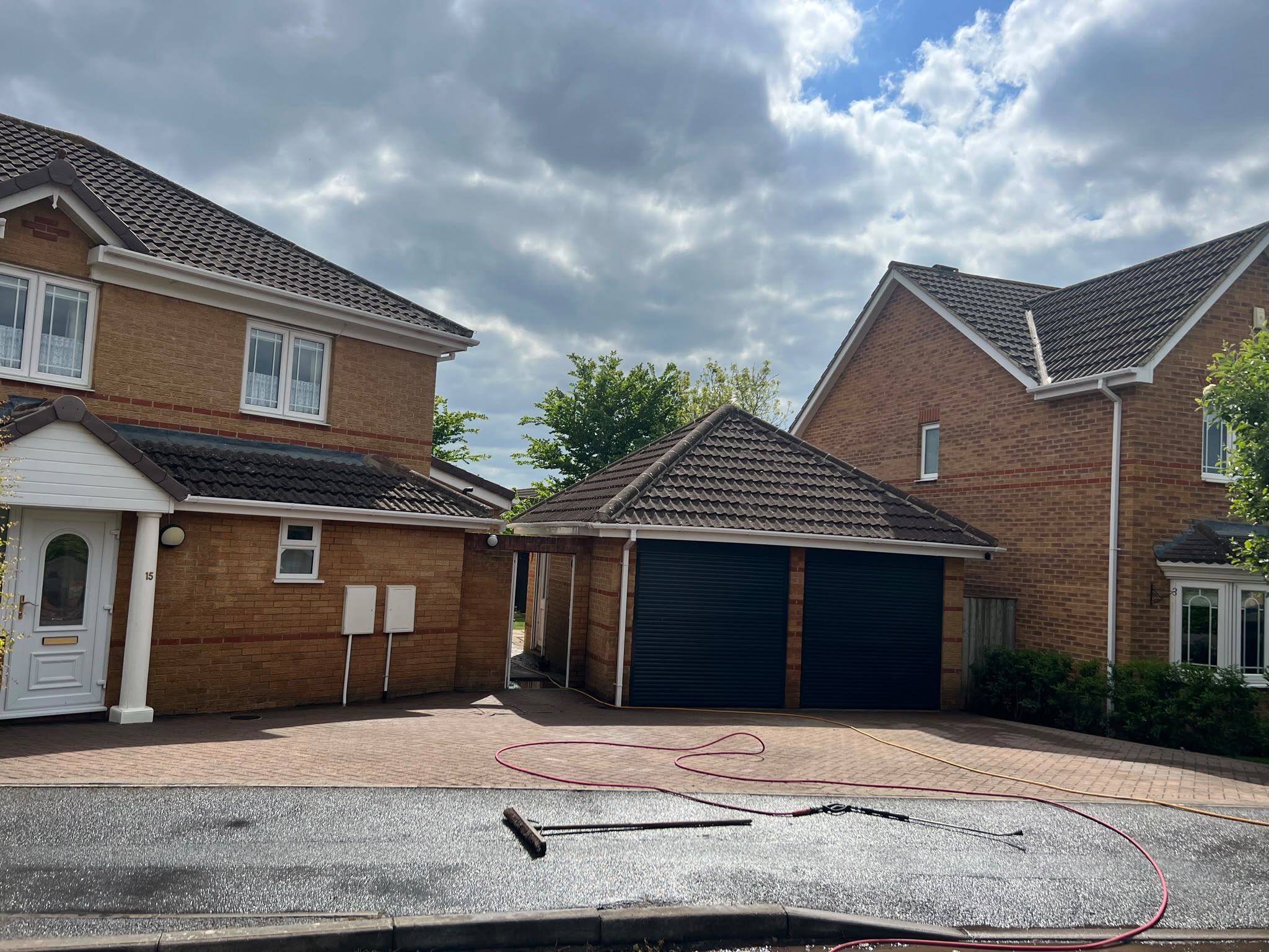 Clean reddish block-paved driveway with hoses and a squeegee on wet tarmac in front of brick houses and a garage
