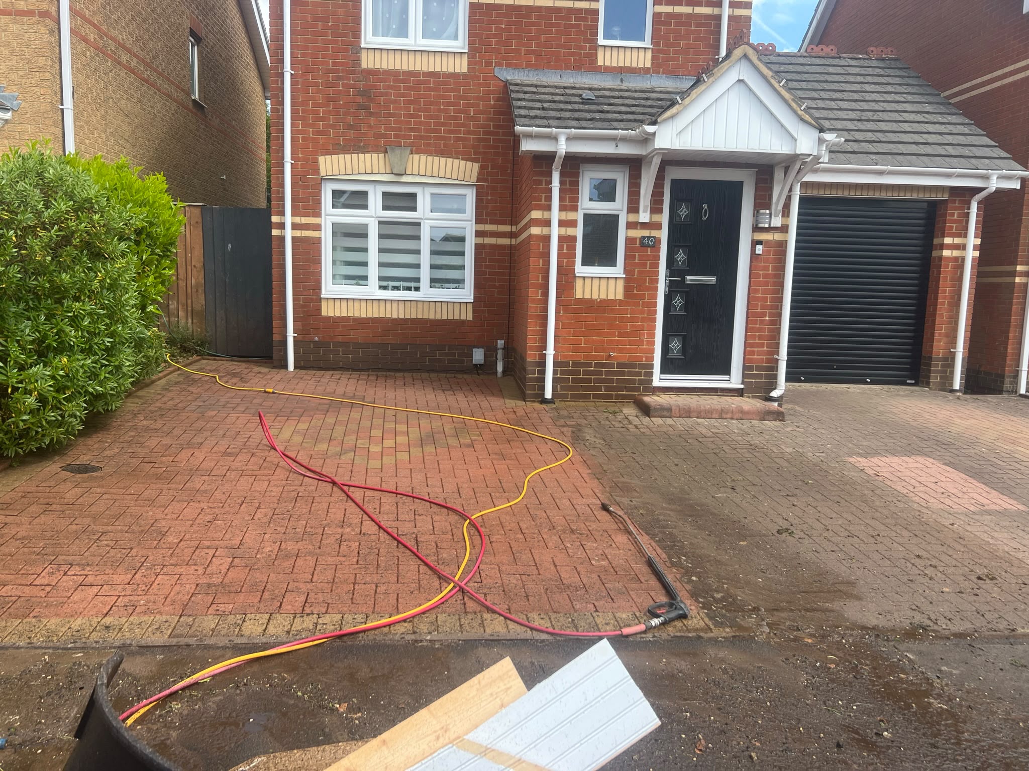 Driveway with bright cleaned paving on one side and dirty paving on the other, wand and hoses in front of a brick house
