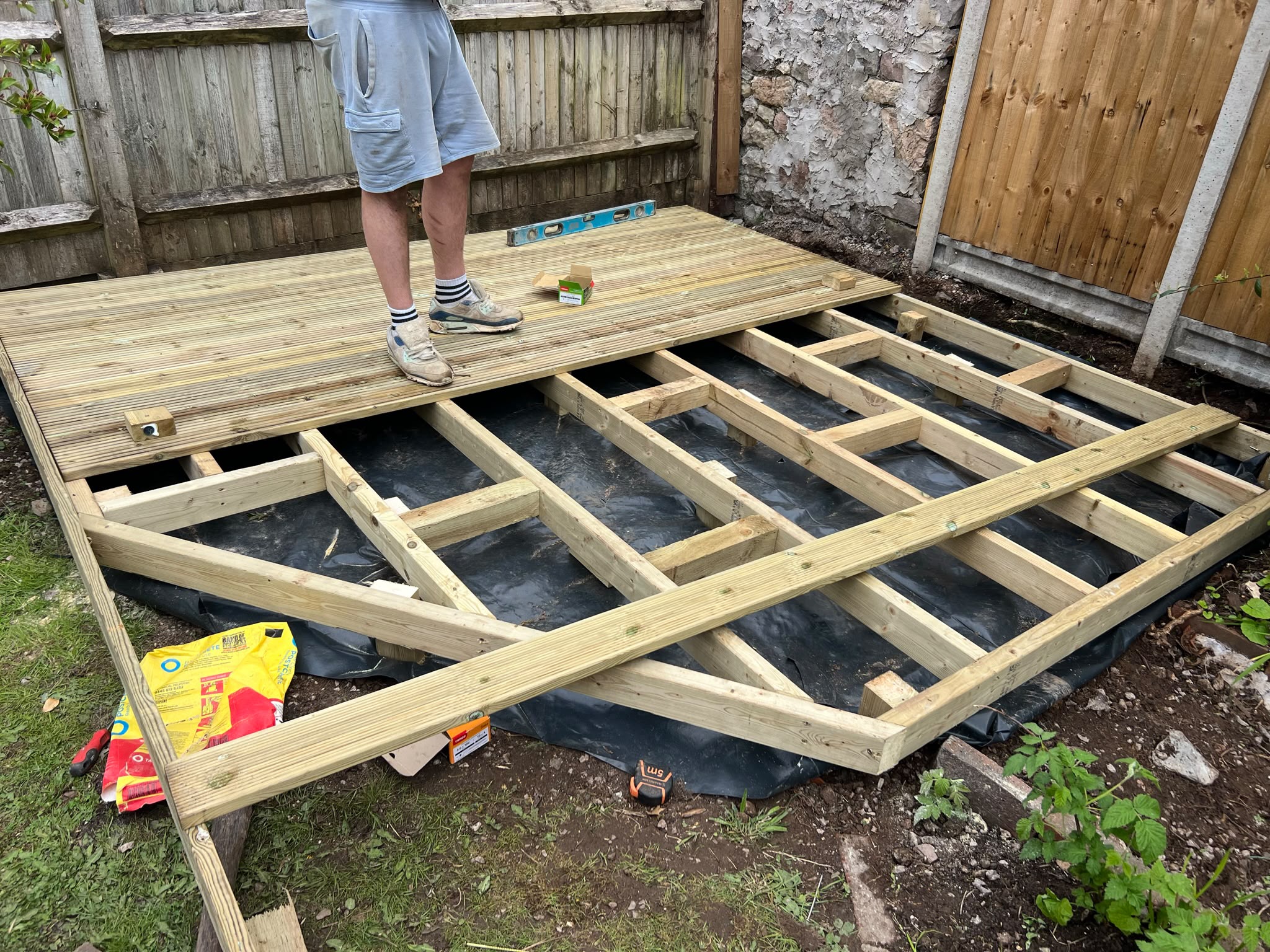 Garden deck under construction with grooved boards over weed membrane, spirit level and screws on the frame