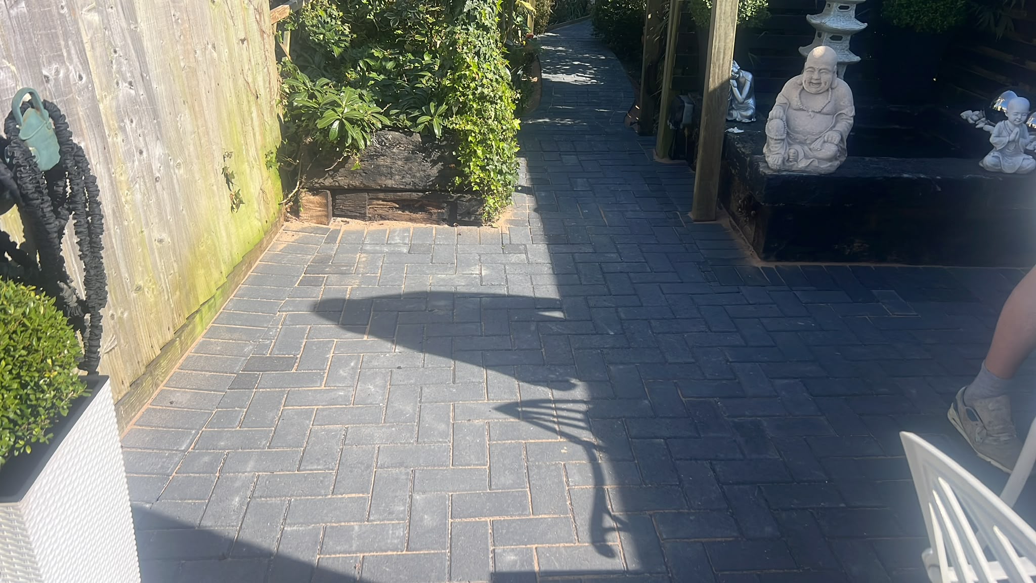 Dark charcoal herringbone block paving patio and path beside timber beds, fencing, and garden statues