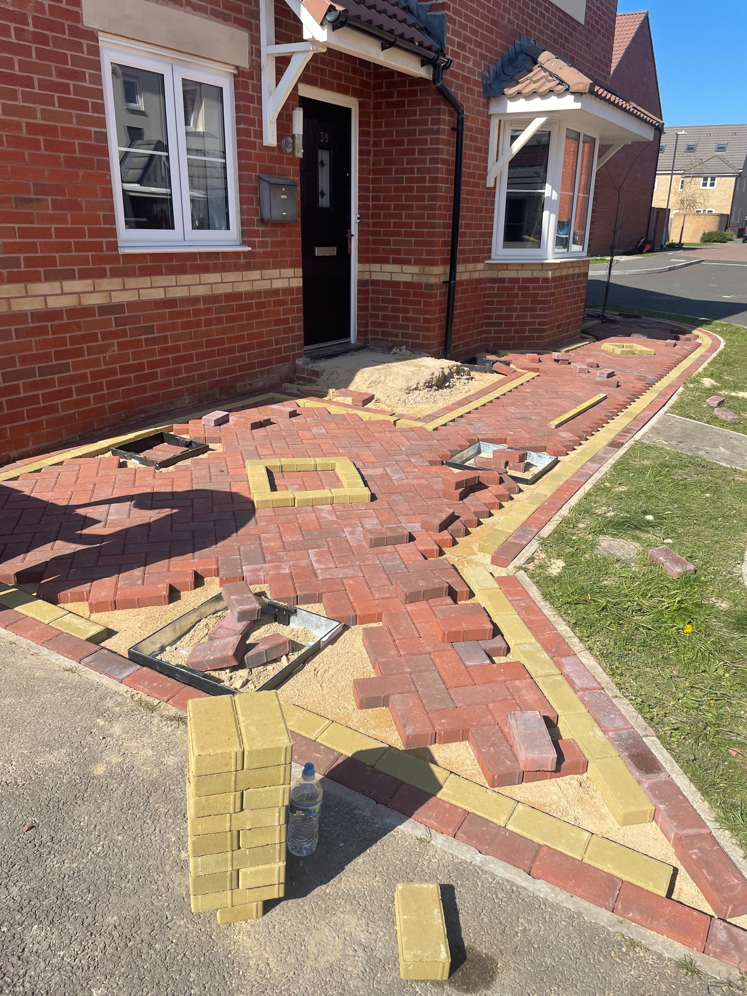 Red and yellow block paving being laid in herringbone on sand in front of a red brick house