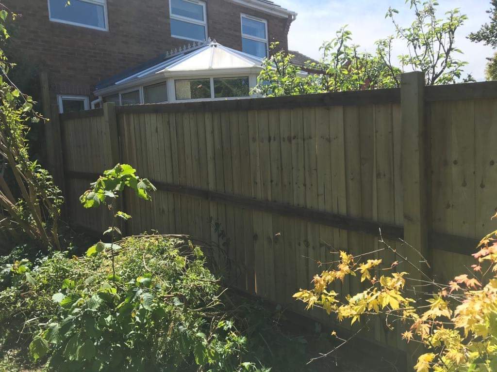 Tall new timber closeboard fence with posts and rails in a garden with shrubs and a brick house beyond