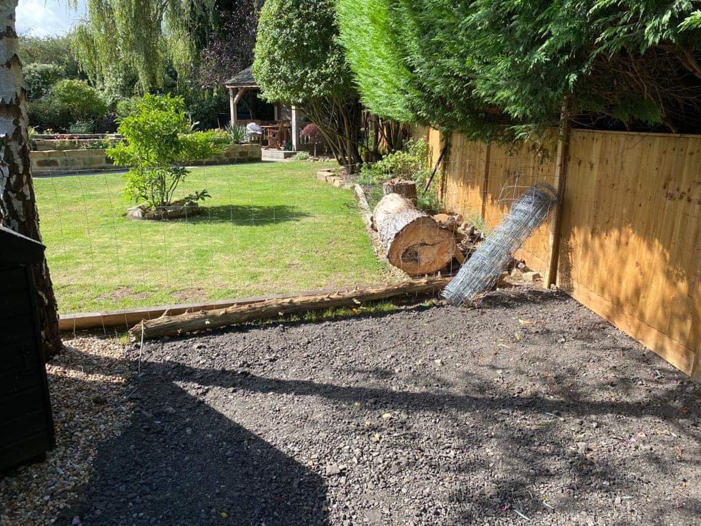 Cleared garden area with dark gravel base, felled tree logs, new wooden fence, and lawn beyond