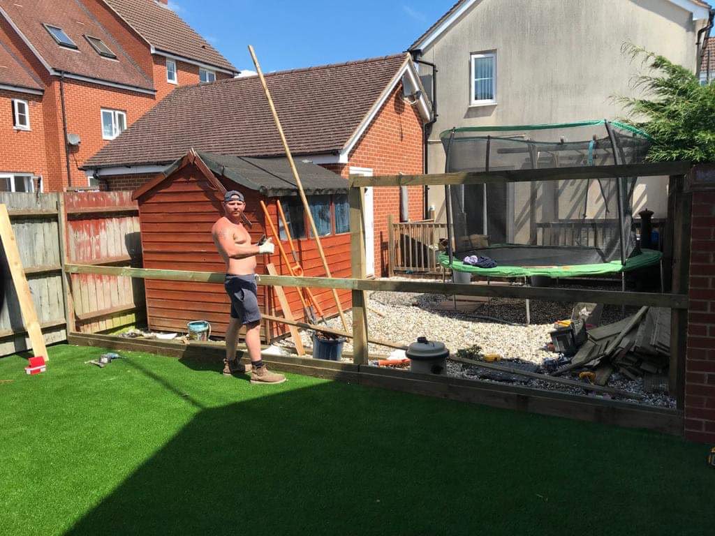 Worker giving thumbs up beside a wooden fence frame of posts and rails in a garden with artificial grass