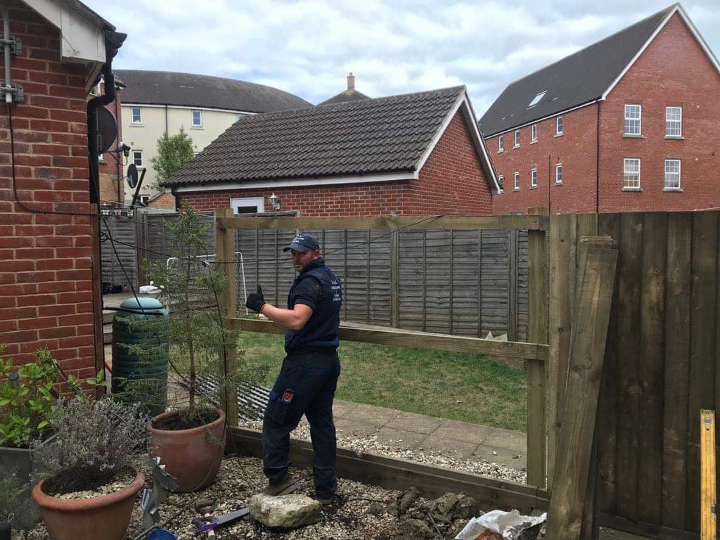Worker in branded workwear giving thumbs up beside posts, rails, and a leaning fence panel in a gravel garden