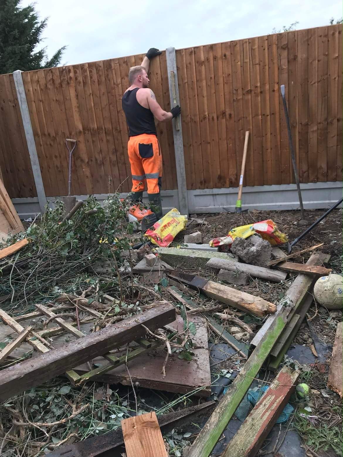 Worker with spirit level checking a concrete fence post beside new reddish featheredge fencing and clearance debris