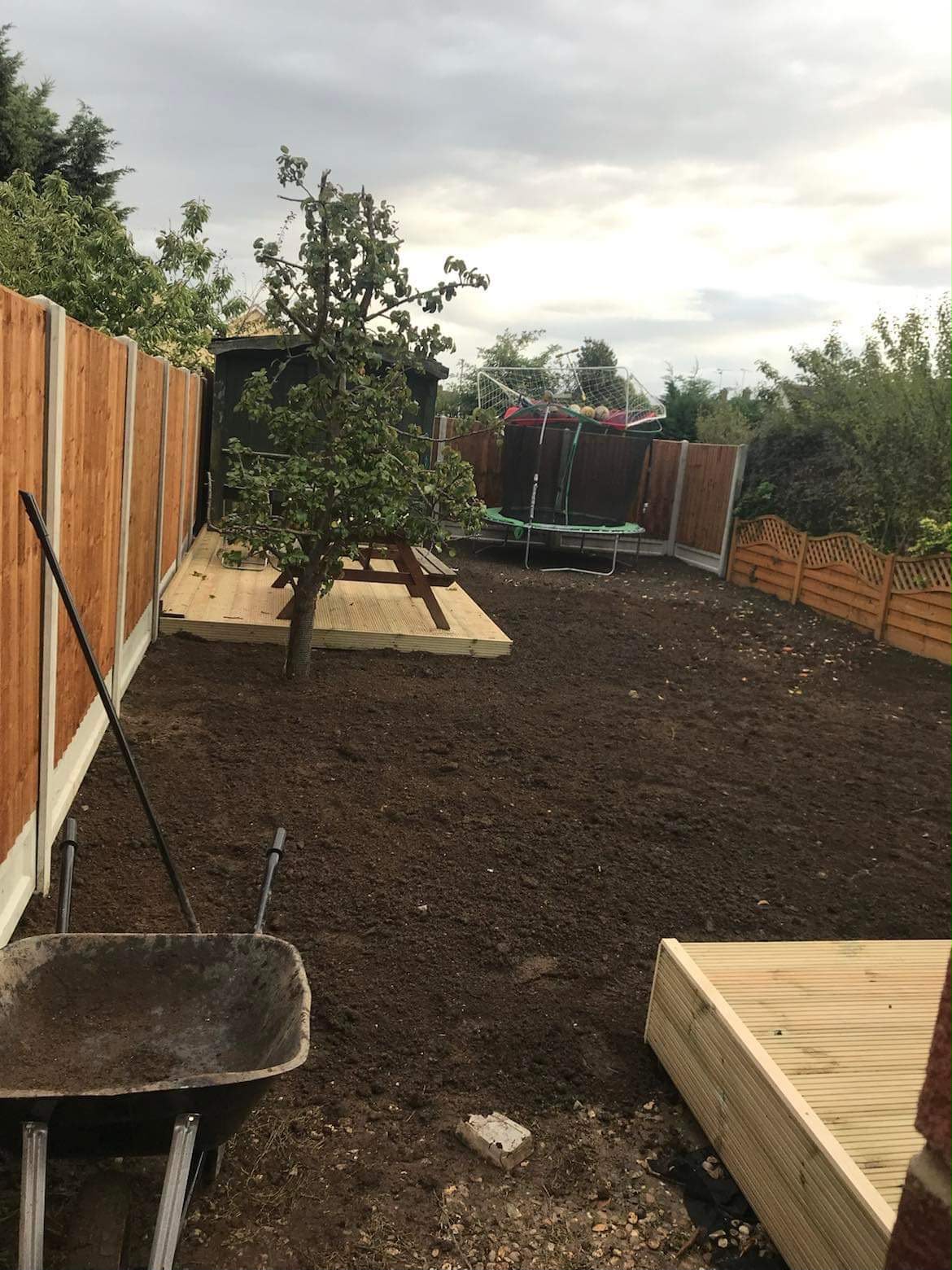 Back garden renovation with freshly turned soil, new timber decking sections, fencing, and a wheelbarrow