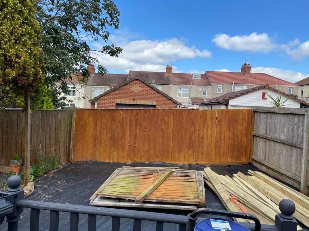 New orange-brown fence panel standing over weed membrane with old panels stacked and timber ready nearby