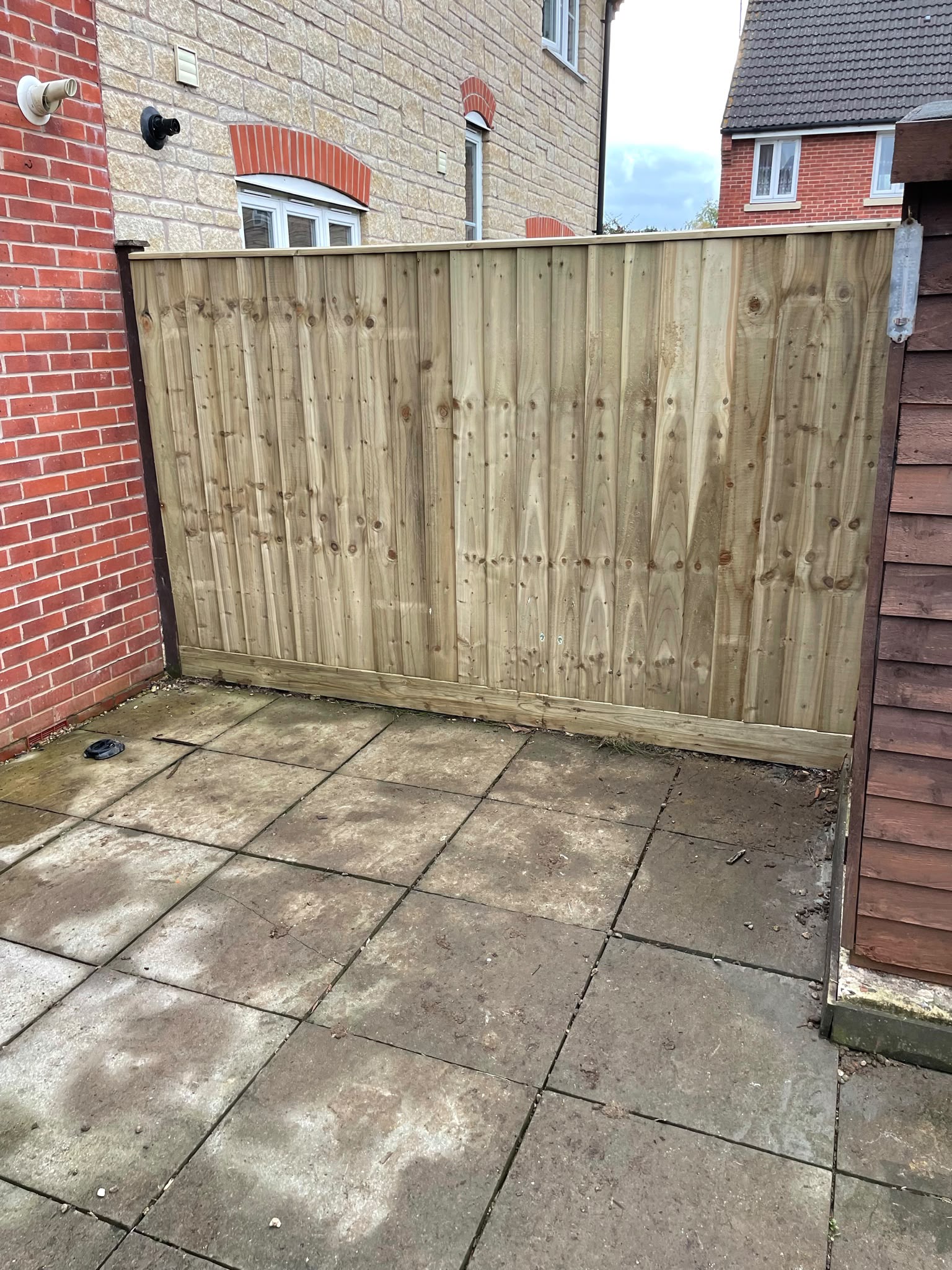 Newly installed light closeboard fence panel with gravel board between a brick house wall and garden shed