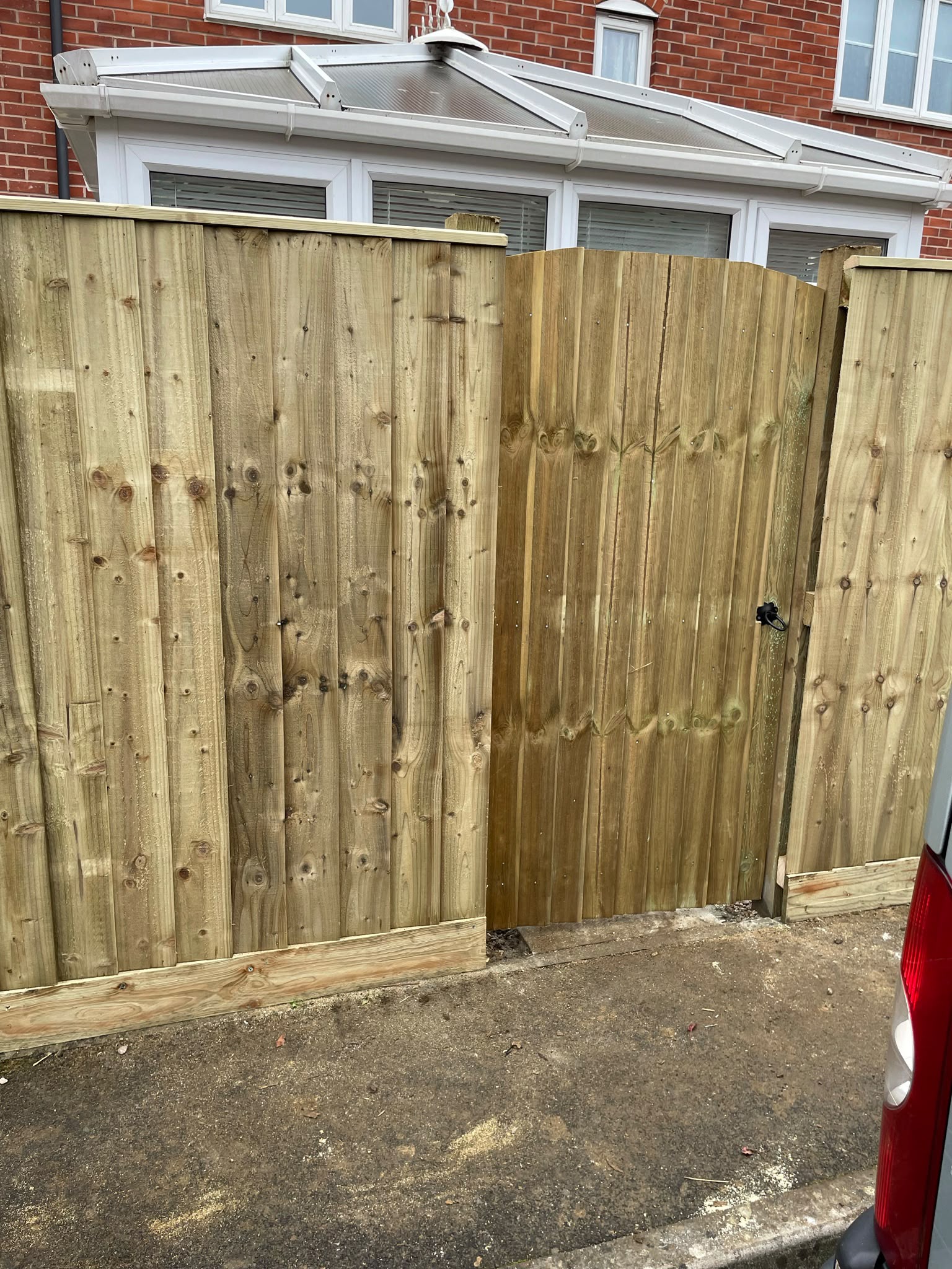 New light timber closeboard fence with matching arched gate and black latch in front of a white conservatory